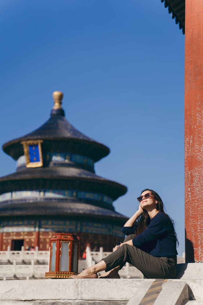 Una mujer joven con gafas de sol se sienta al sol y mira hacia el cielo en los escalones del Templo del Cielo en Beijing, con el distintivo pabellón circular de fondo.