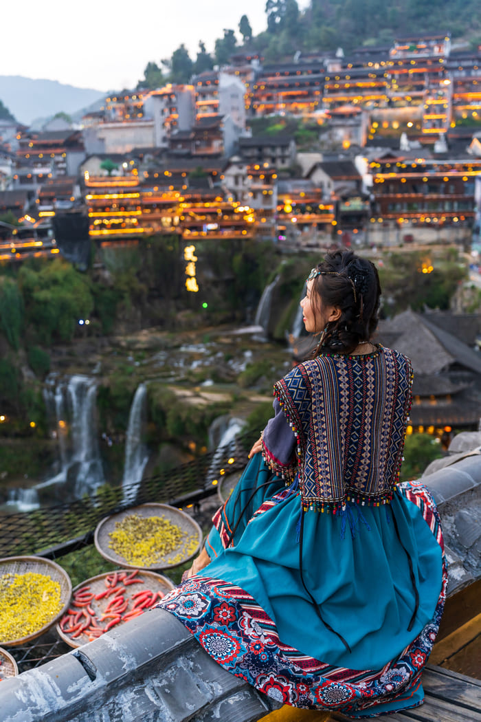 Mujer con vestido tradicional contempla las cascadas y las casas iluminadas del antiguo pueblo de Furong en Hunan, China, durante el atardecer.