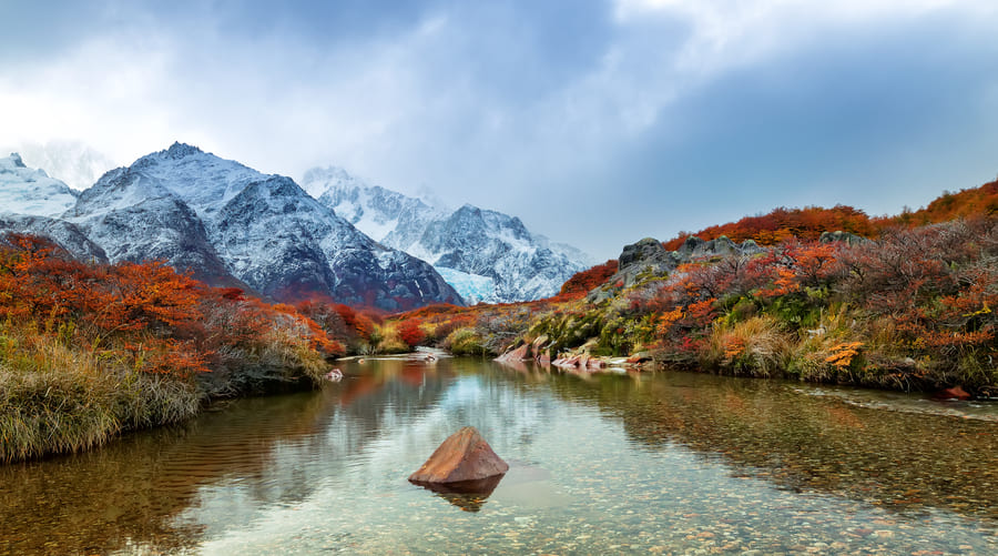 Paisaje de otoño en la Patagonia argentina con un río cristalino en primer plano y montañas nevadas del Monte Fitz Roy bajo un cielo nublado.