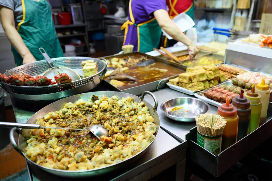 Puesto de comida callejera tradicional en un mercado nocturno asiático con bandejas de marisco guisado, tofu frito y brochetas variadas.
