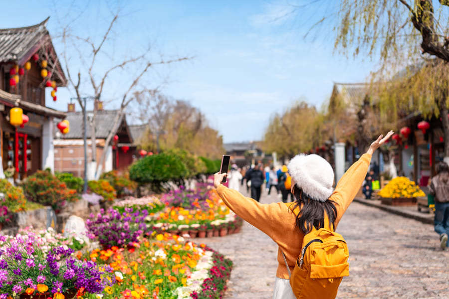 Una turista con mochila amarilla y sombrero blanco se toma una selfie en una calle florida de la Ciudad Antigua de Lijiang en Yunnan, China.