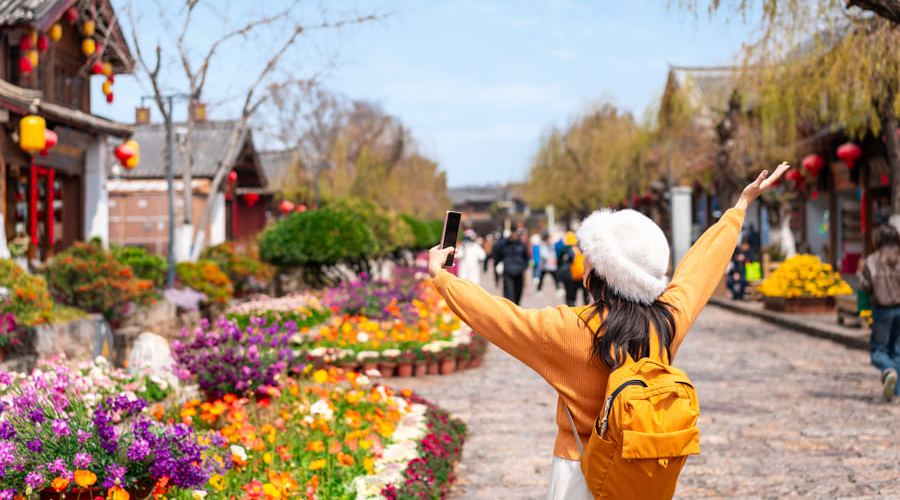 Una turista con mochila amarilla y sombrero blanco se toma una selfie en una calle florida de la Ciudad Antigua de Lijiang en Yunnan, China.