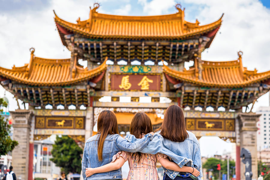 Tres amigas abrazadas de espaldas contemplan el histórico Arco de la Jinma Biji en una plaza céntrica de Kunming, Yunnan, China.