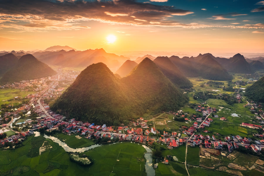 Una vista aérea panorámica muestra un pintoresco paisaje rural en Yangshuo al atardecer, donde un pequeño pueblo con edificios de techos naranjas y blancos se extiende a lo largo de un río serpenteante en un valle verde